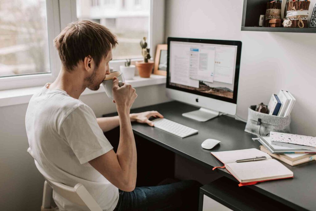 A remote worker sits in front of their computer in their home office, sipping coffee and checking their email. 